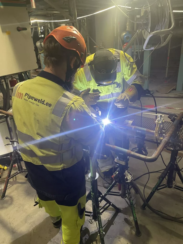 Två svetsare monterar en komponent till en ångpanna på tripoder inom industrirör.