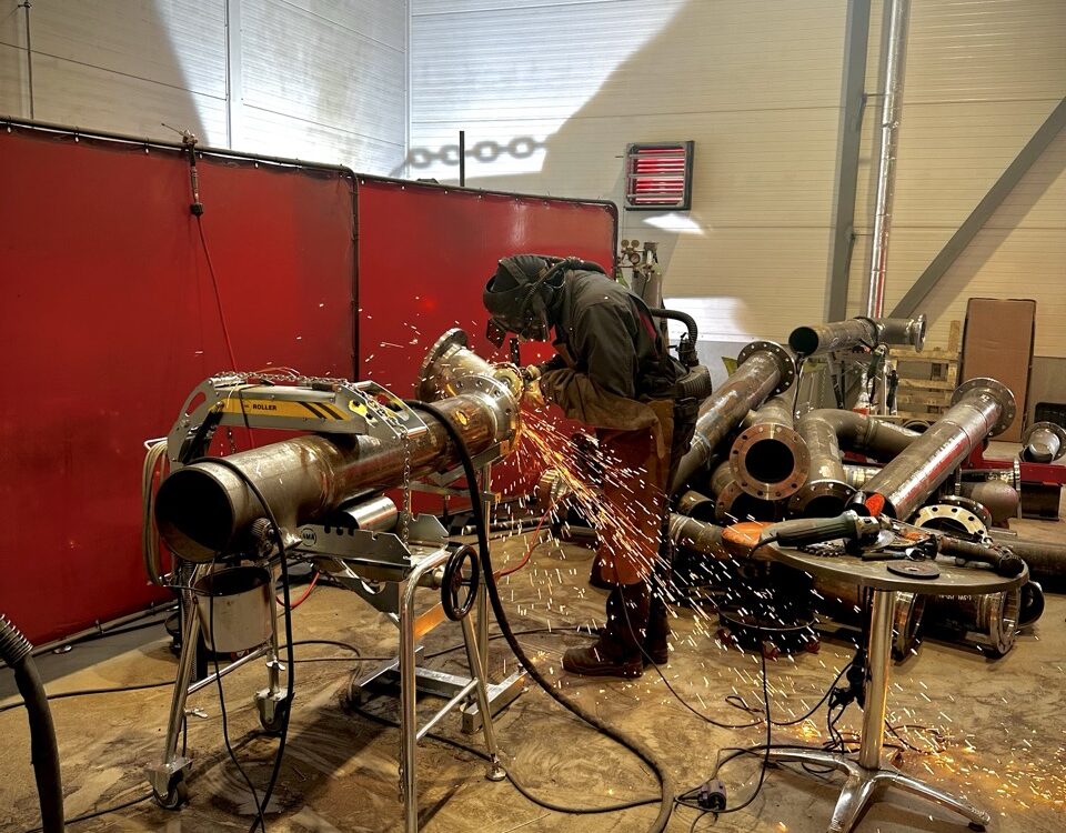 A welder working on a pipe in a fabrication workshop, with multiple prefabricated pipe sections neatly stacked on the right, ready for further processing and assembly.